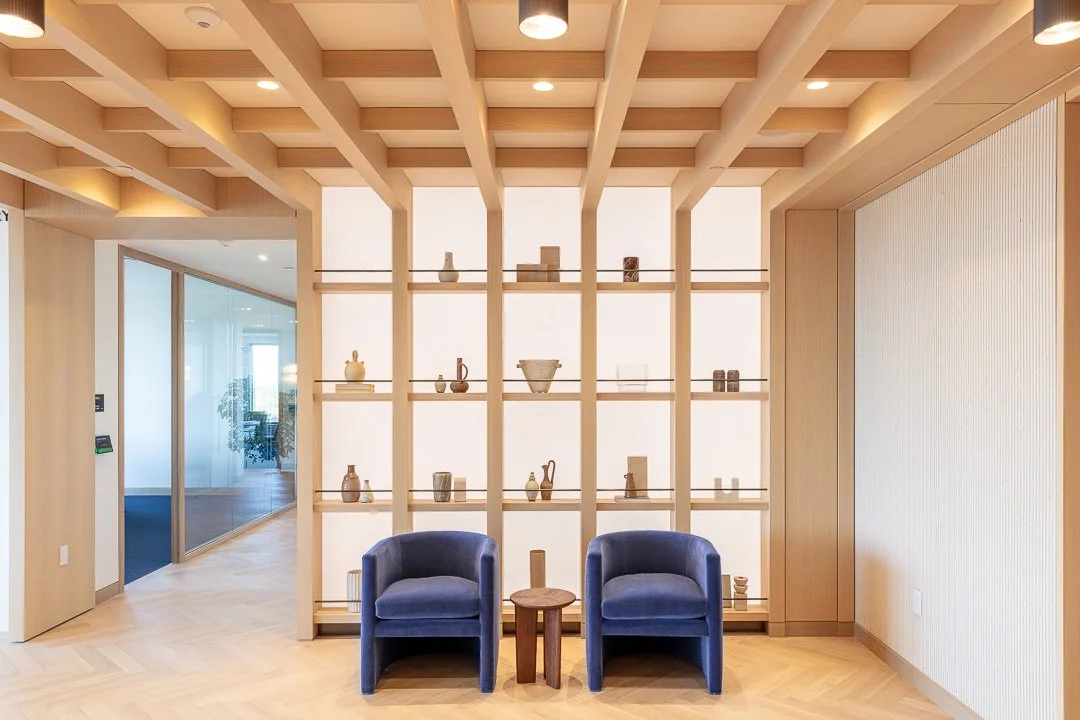 Two blue armchairs with a small wooden table between them in front of a decorative wooden grid with ceramic vases and objects on shelves, in a modern, minimalist room.