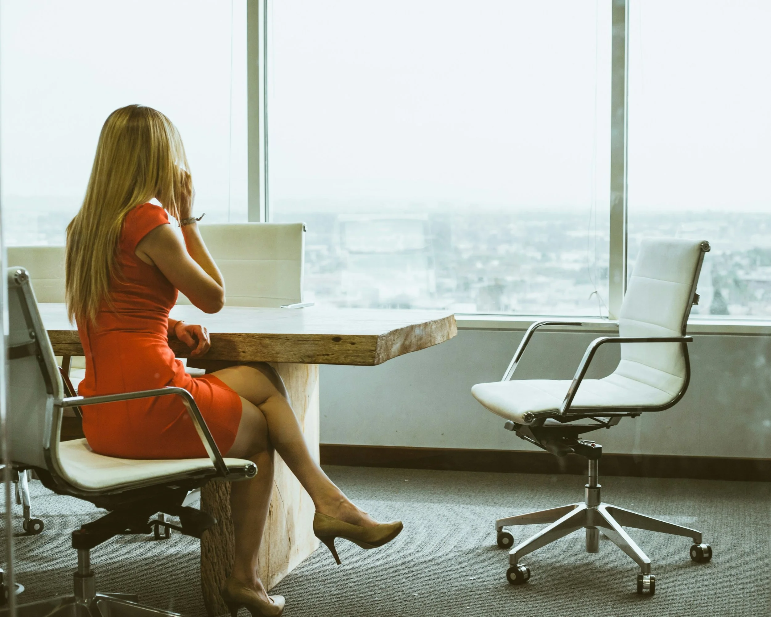 A woman with long red hair wearing a red dress and high heels sitting in an empty office with large windows overlooking a cityscape.