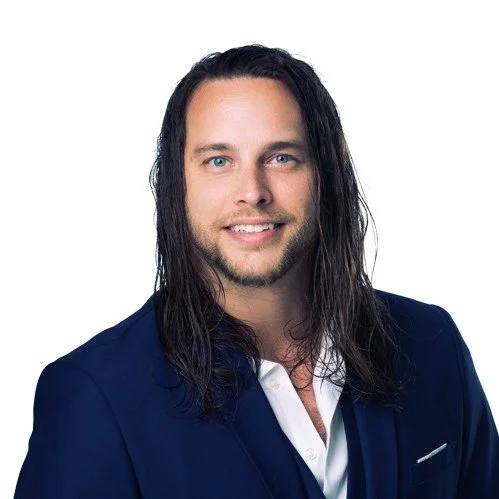 Portrait of a man with long dark hair, blue eyes, smiling, wearing a dark blue blazer and white shirt against a white background.