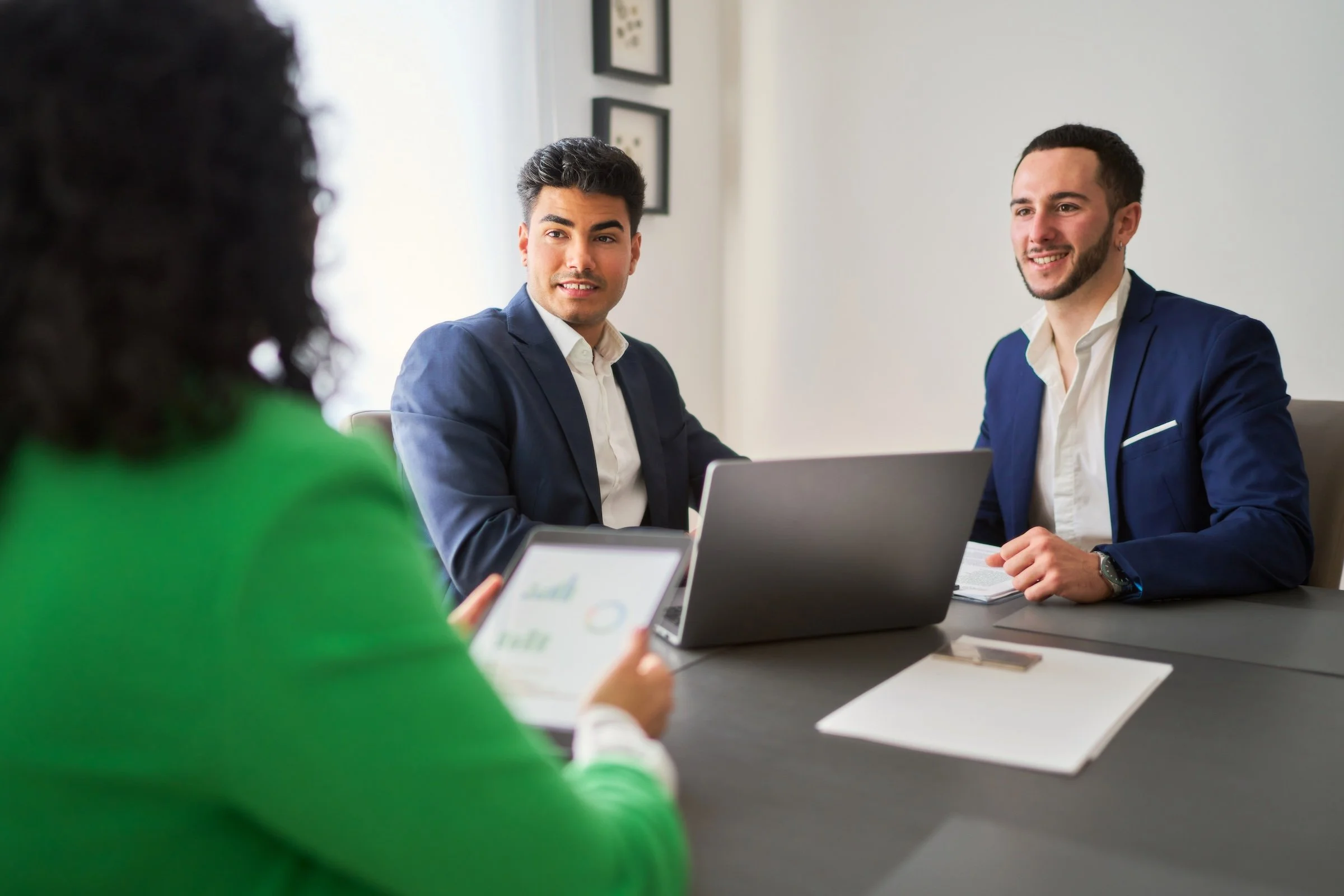 Three people in business attire having a meeting in a conference room. Two men are facing the camera, smiling, while a woman with curly dark hair in a green blazer is seen from behind, holding a tablet showing graphs.