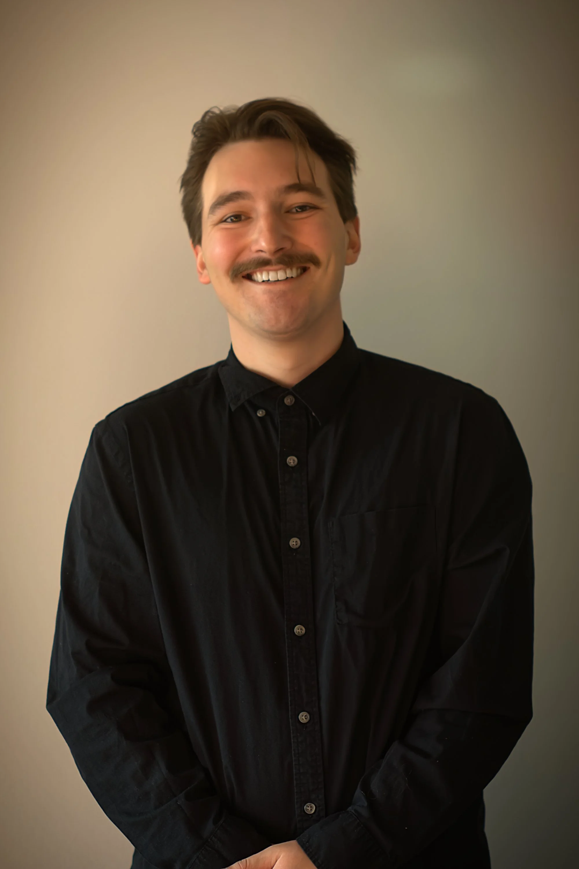 A young man with light skin, brown hair, and a mustache, smiling and wearing a black button-up shirt, standing against a plain light-colored wall.