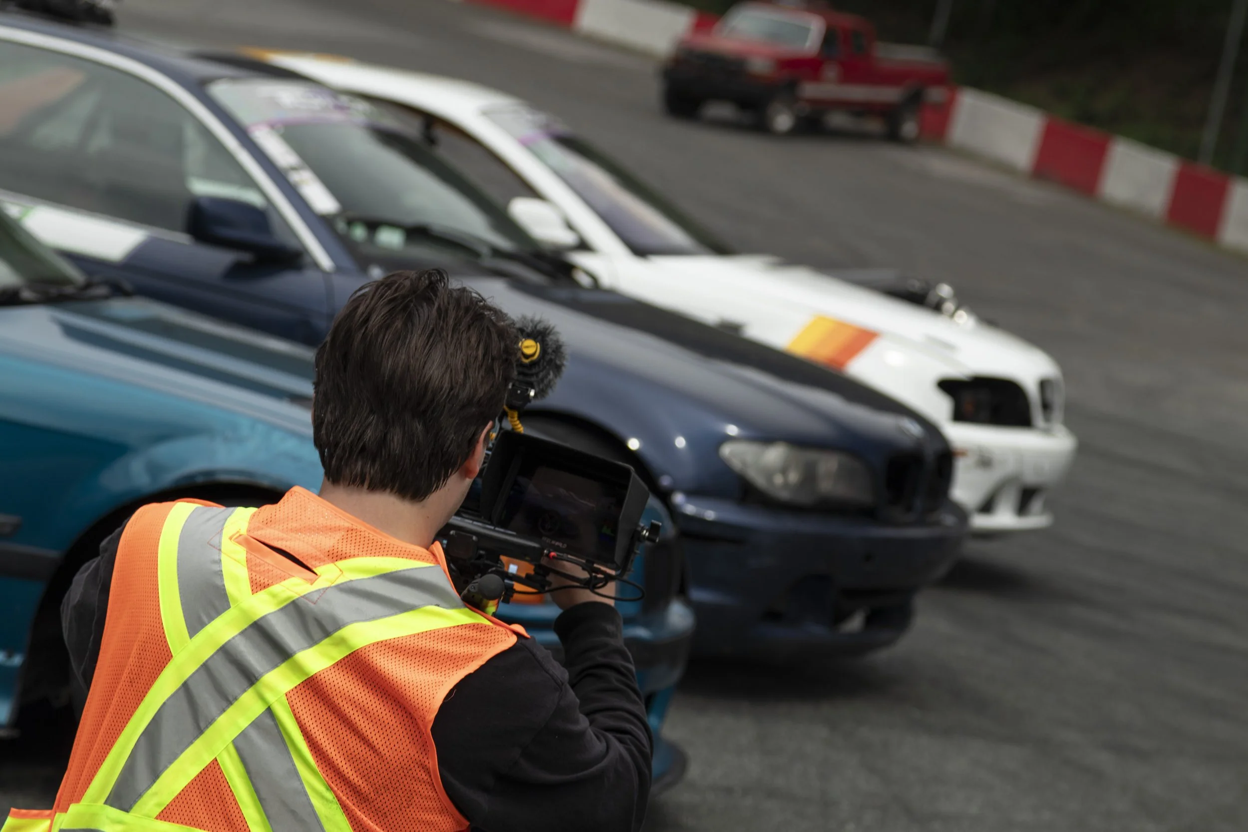 A man in a high-visibility vest taking photographs of a car accident scene on a race track, with damaged cars in the background.