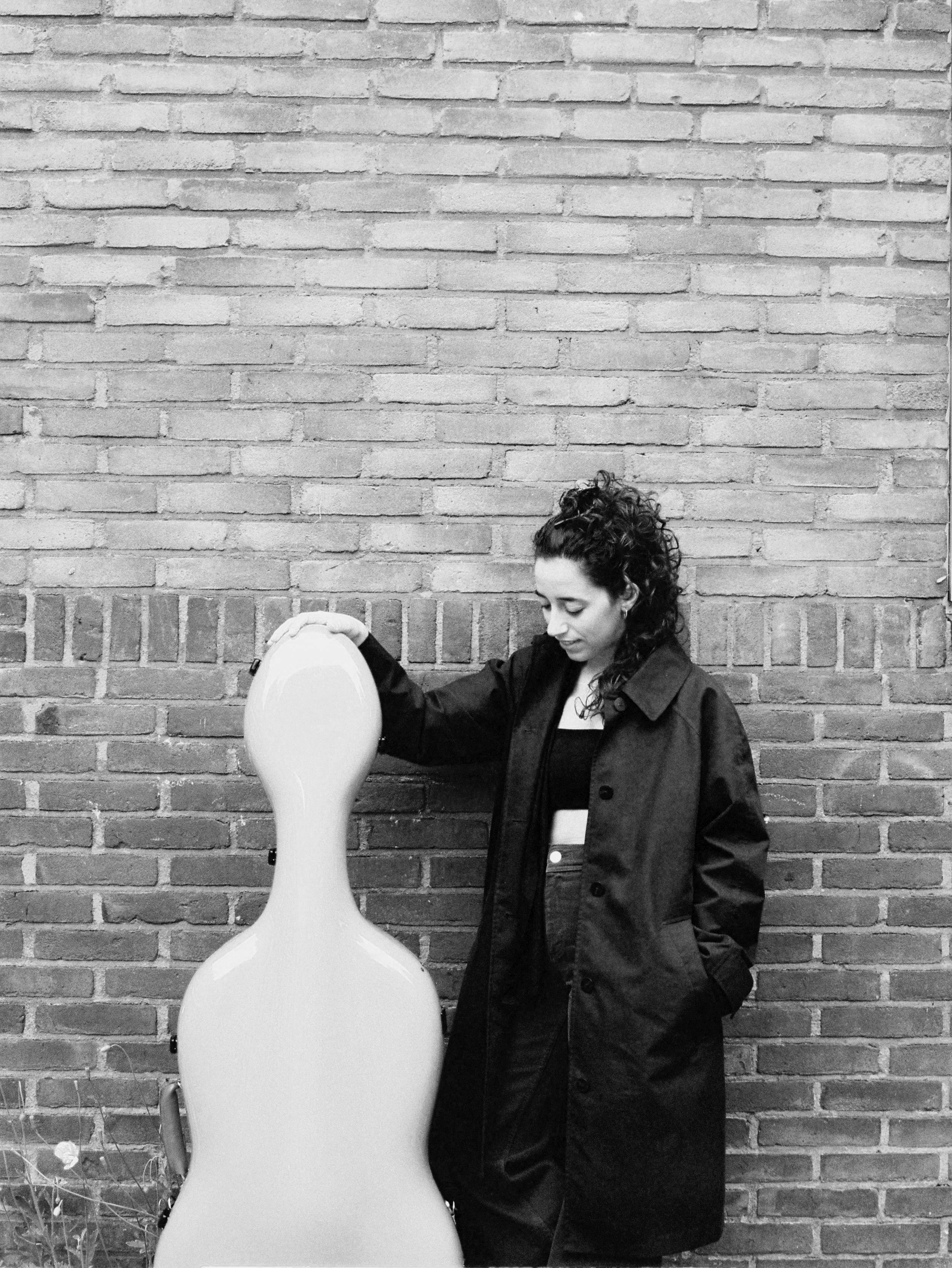 A woman with curly hair wearing a black coat, standing against a brick wall, holding a white balloon.