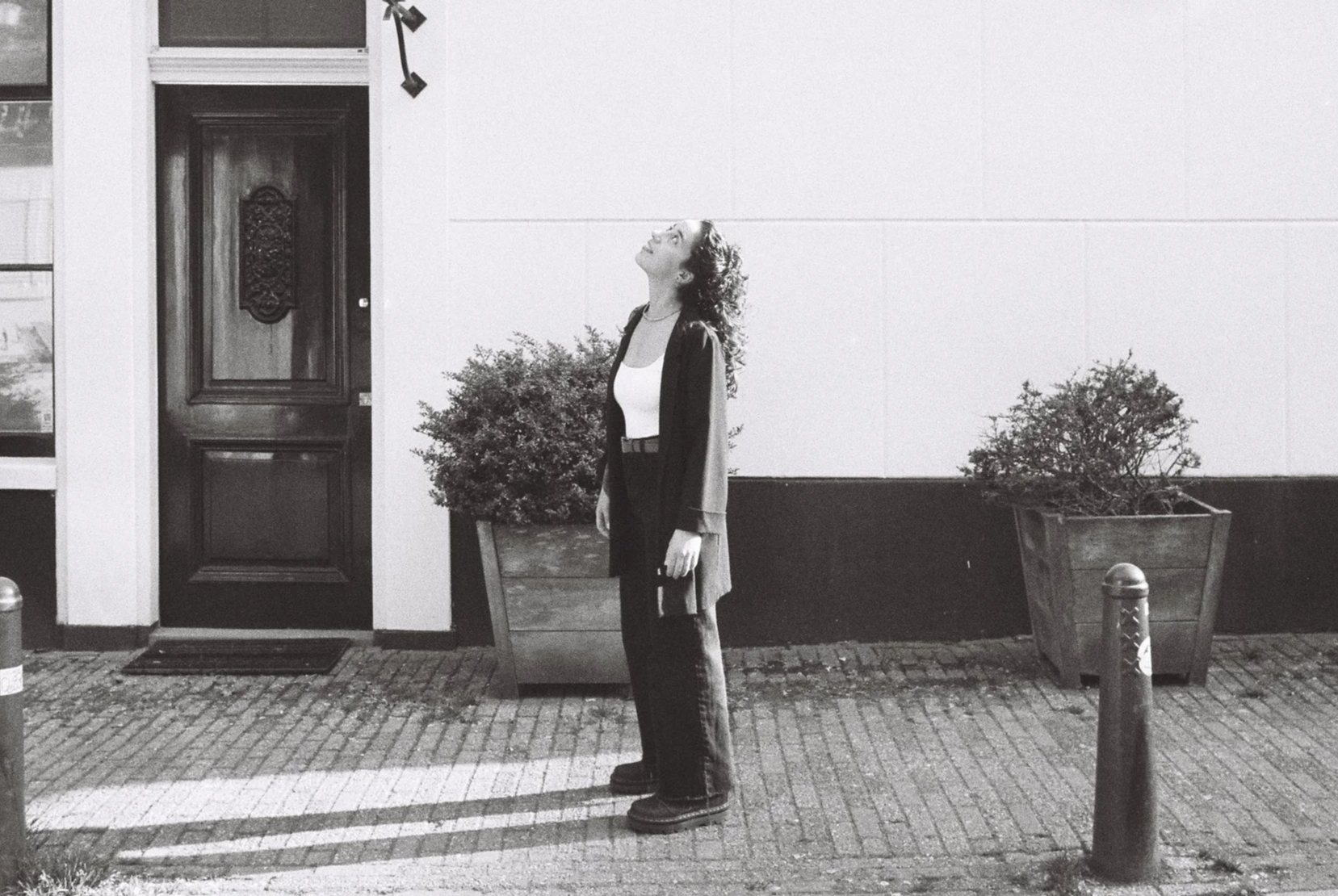 A woman standing on a sidewalk at night, looking up with her eyes closed, wearing a light-colored top, dark pants, and a dark coat. She is holding a small purse in her right hand. There are large potted plants behind her, a wooden door to her left, and a metal bollard in front of her. The scene is in black and white.