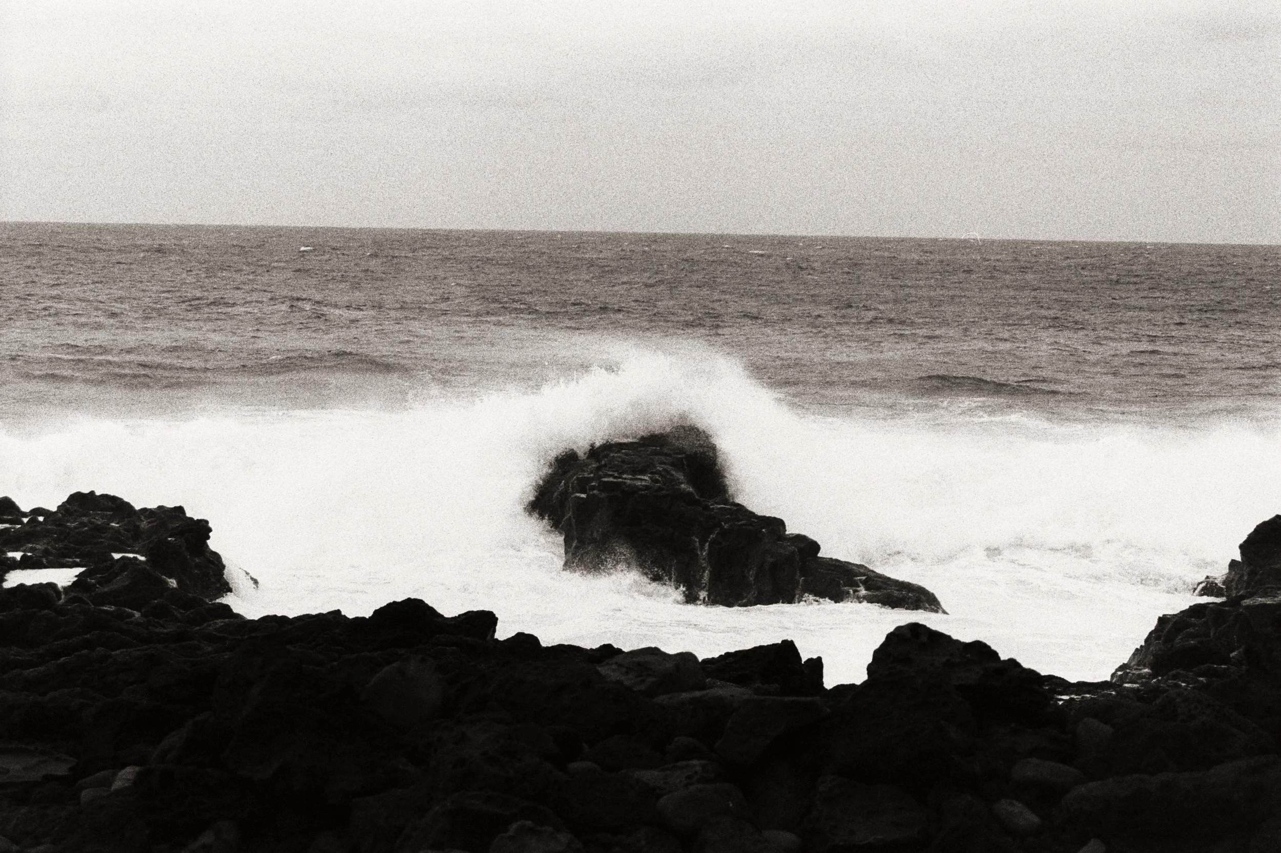 Black rocks along the coast with waves crashing against them and a vast ocean in the background under a cloudy sky.