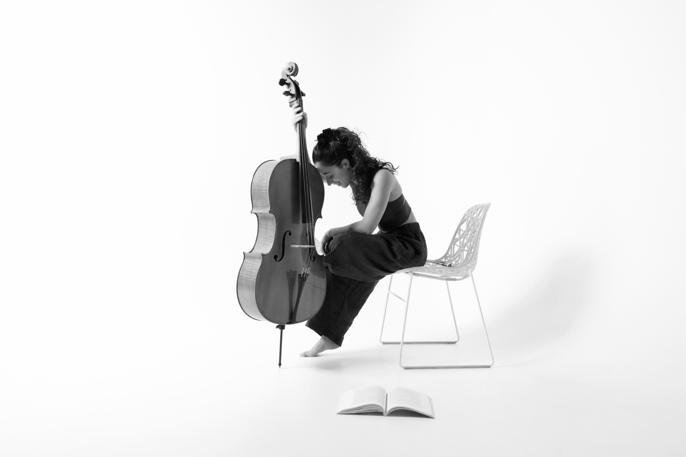 A woman sits with her head bowed and eyes closed, resting her forehead against a cello. She is seated on a white chair, with an open book on the floor in front of her and a music stand behind her. The scene is minimalistic with a plain white background, and the image is in black and white.