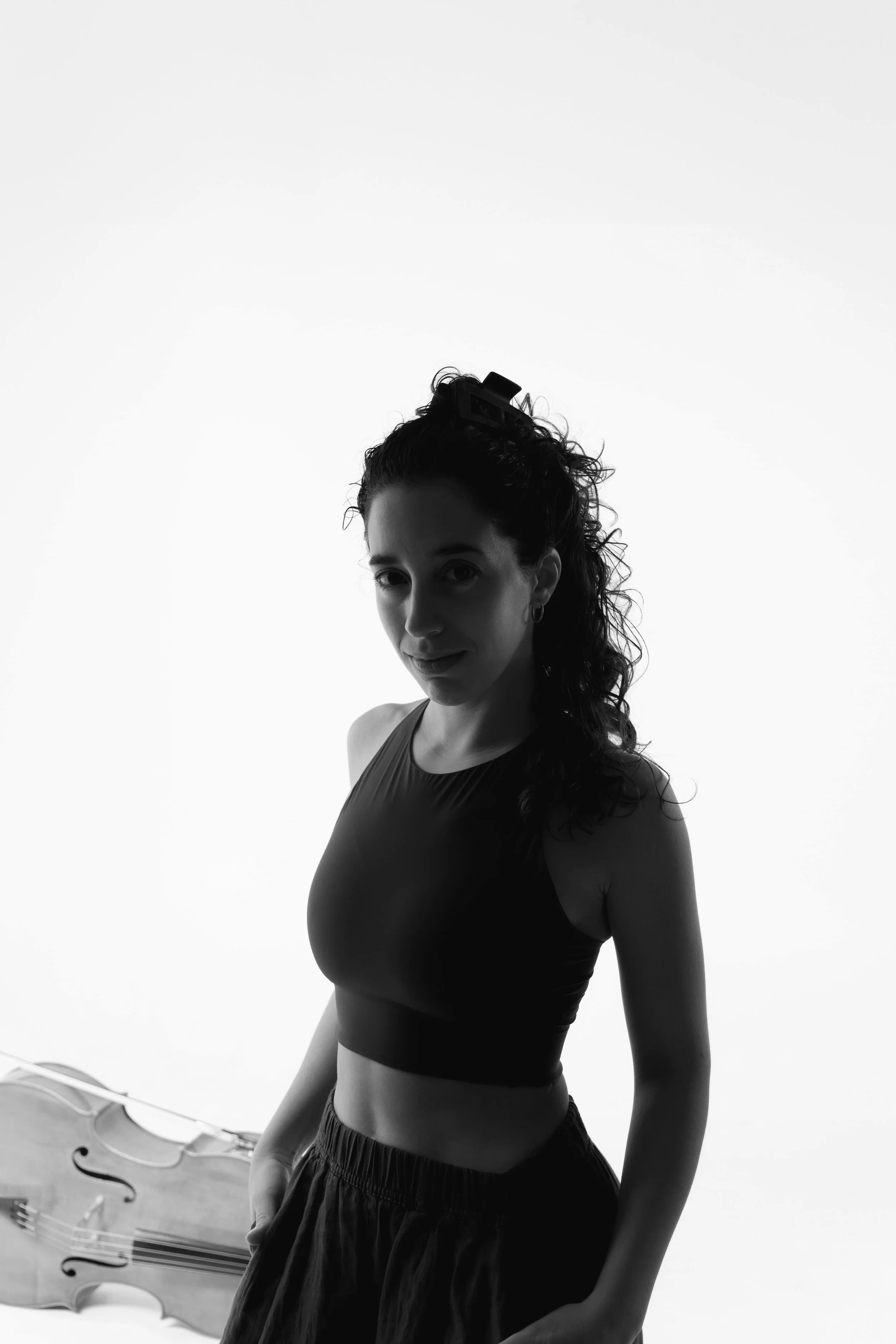 A black-and-white photo of a young woman with long curly hair, wearing a sports bra and shorts, standing indoors near a wall with a violin on a stand behind her.