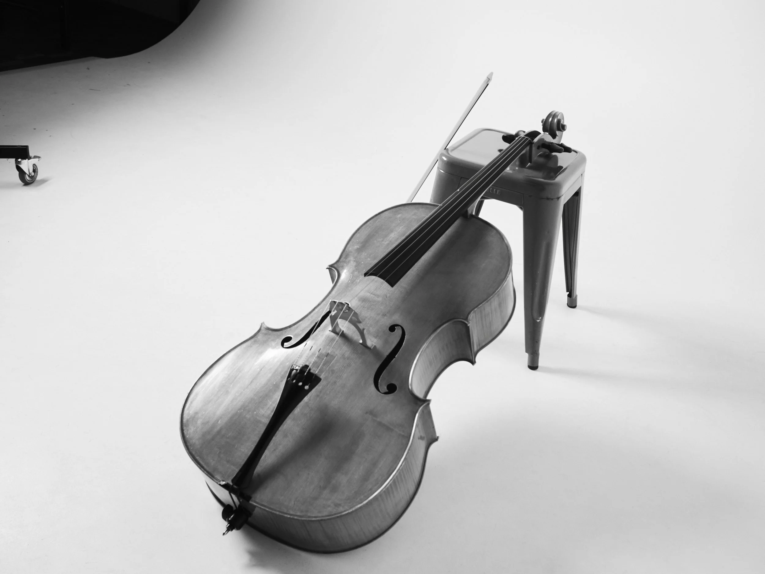 A close-up of a wooden cello resting on a small stand with a bow placed on top of it, on a white surface.