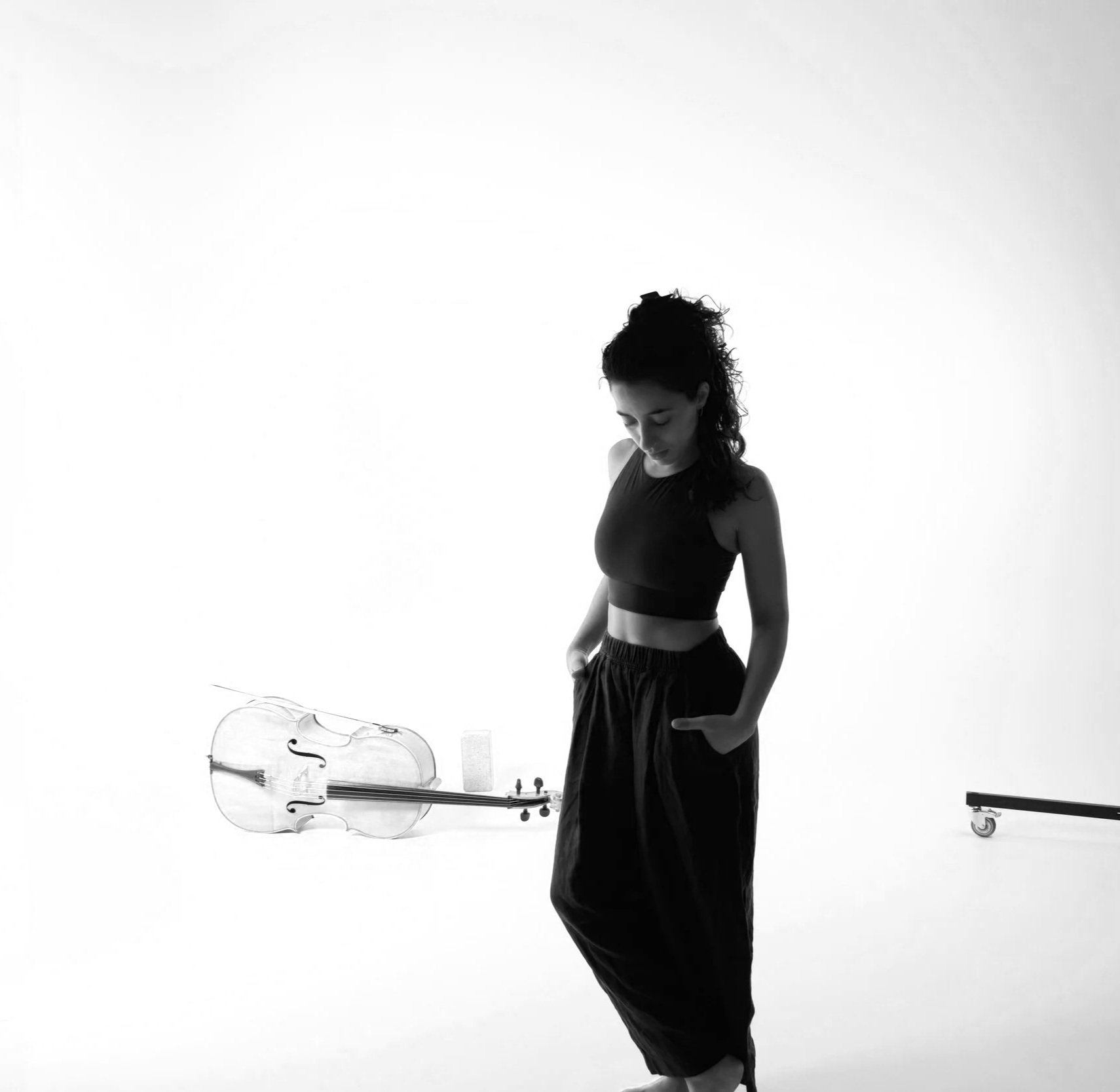 A woman standing in a photography studio with a white background, wearing a black sports bra and black pants. She has dark, curly hair and is looking down. A transparent violin and a bow are on the left side of the image, with a small object and a rolling stand on the right.