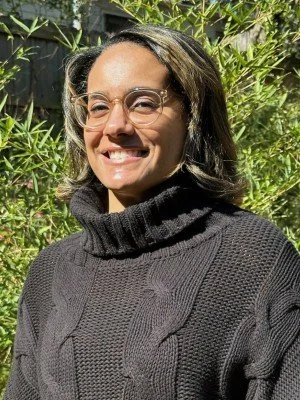 Woman with glasses smiling outdoors, wearing a black turtleneck sweater, surrounded by green foliage.