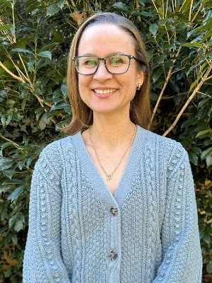 A woman with glasses and shoulder-length brown hair smiling outdoors, wearing a light blue sweater and a necklace, standing in front of green bushes.