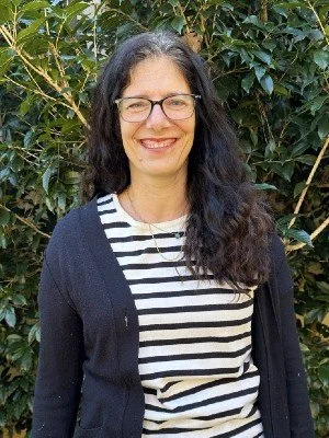 A woman with long dark hair and glasses smiling outdoors, standing in front of green leafy bushes, wearing a striped shirt and dark cardigan.