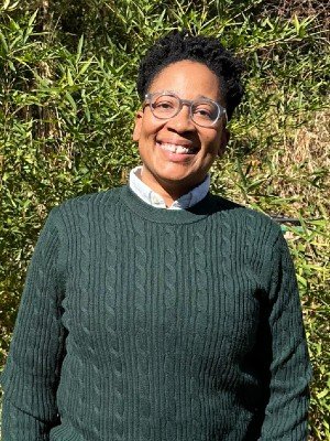 Woman smiling outdoors, wearing glasses and a dark green cable-knit sweater, with greenery in the background.