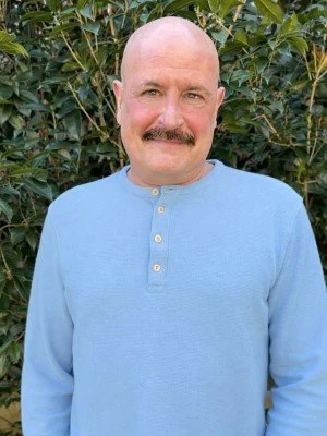A bald man with a mustache wearing a light blue long-sleeve shirt, standing outdoors in front of green foliage.
