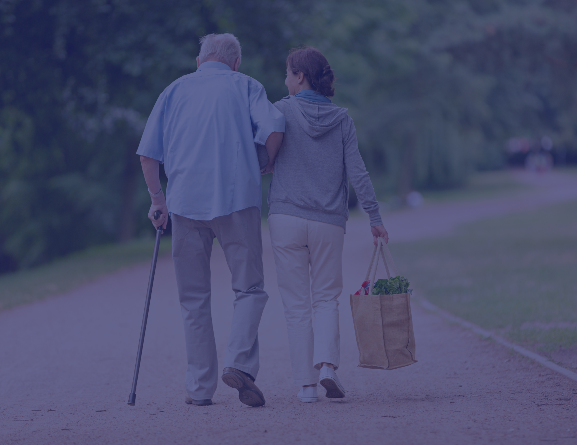 Older man and woman walking together on a park pathway, with the man using a cane and the woman carrying a grocery bag, surrounded by greenery.