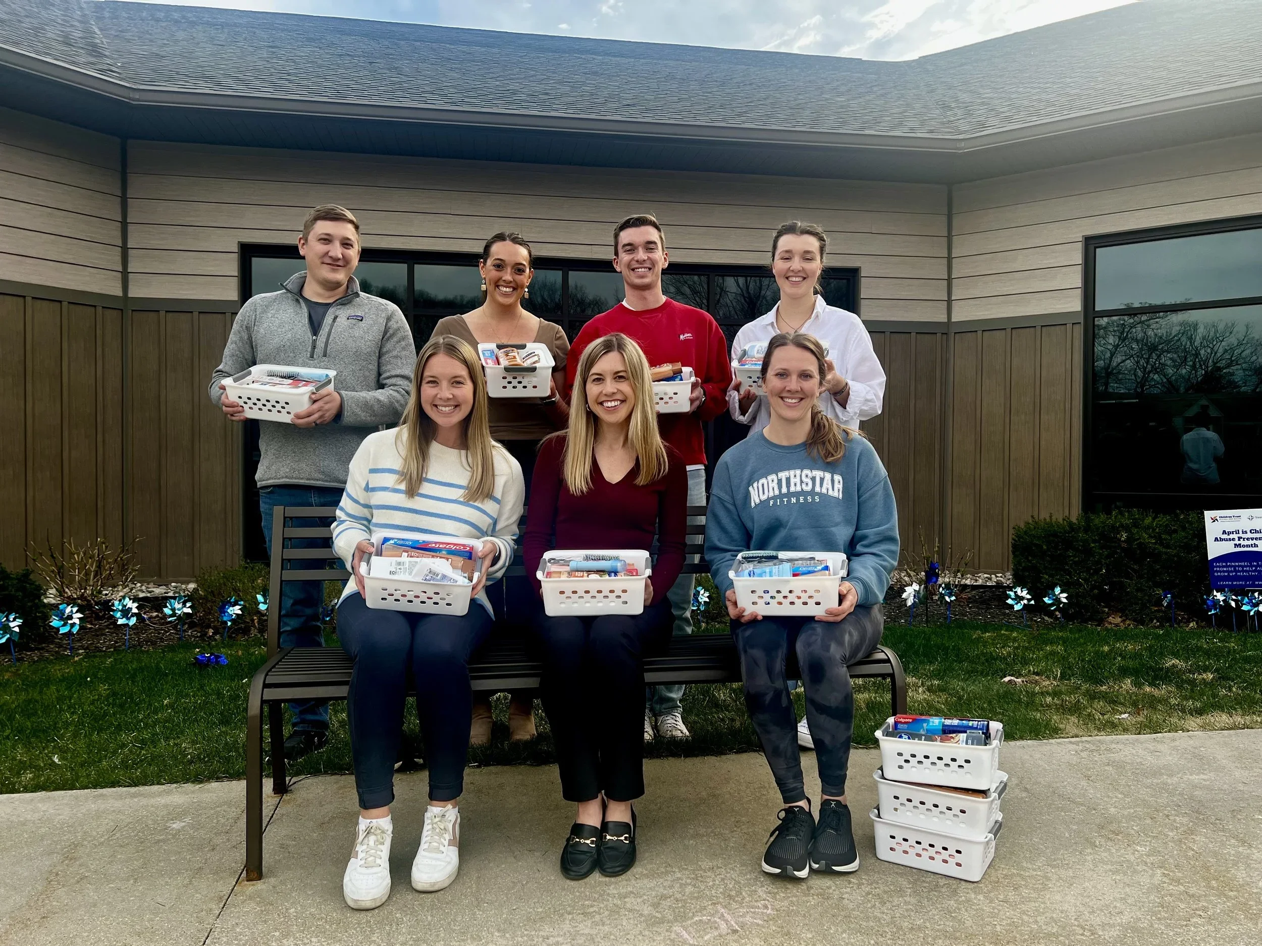 Group of eight smiling young adults holding white baskets with snacks and supplies, standing and sitting in front of a modern house with a lawn and garden, during daytime.
