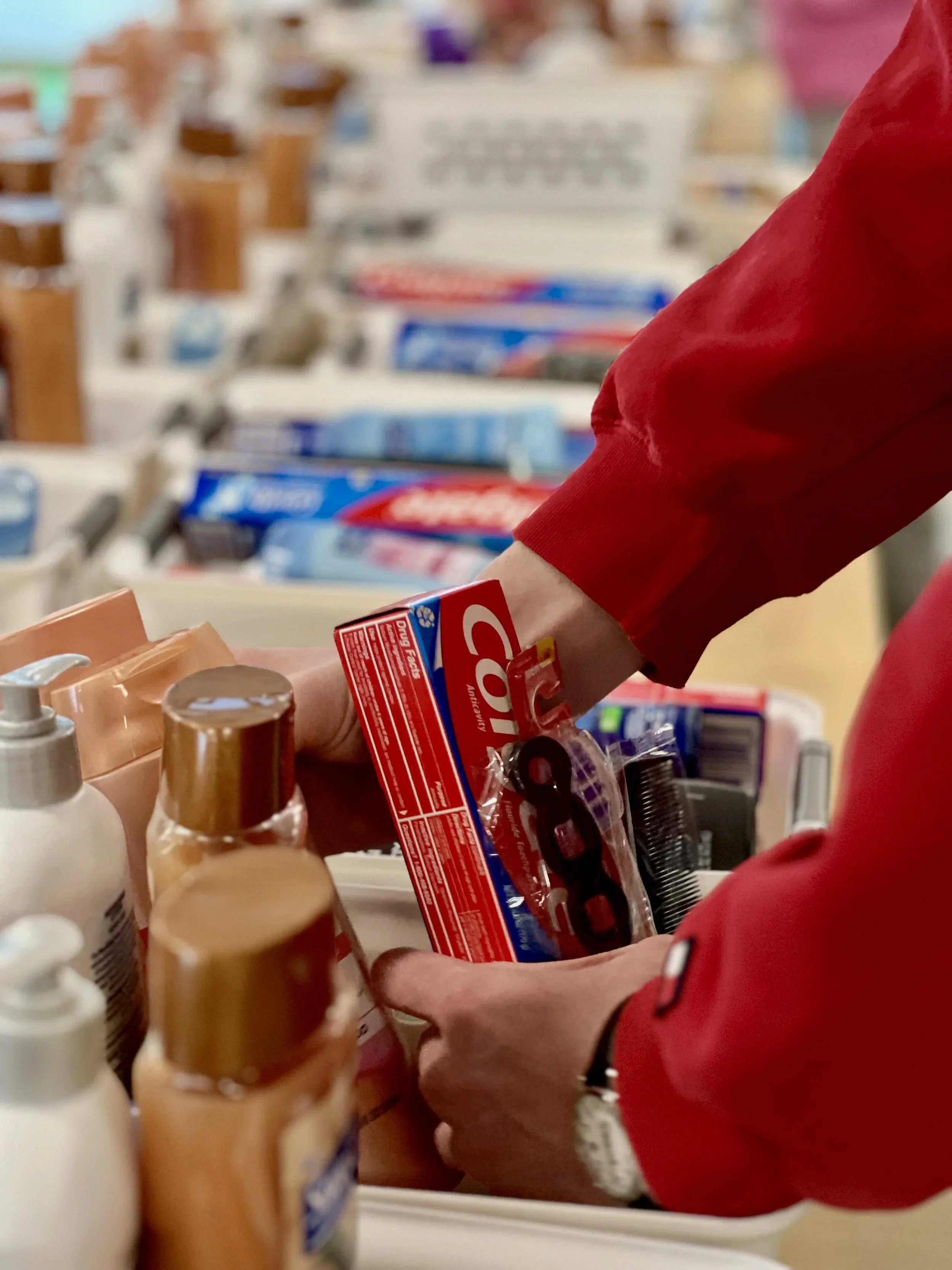 A person wearing a red long sleeve shirt shopping in a store, holding toothpaste and other toiletries near a display of various toiletry items.