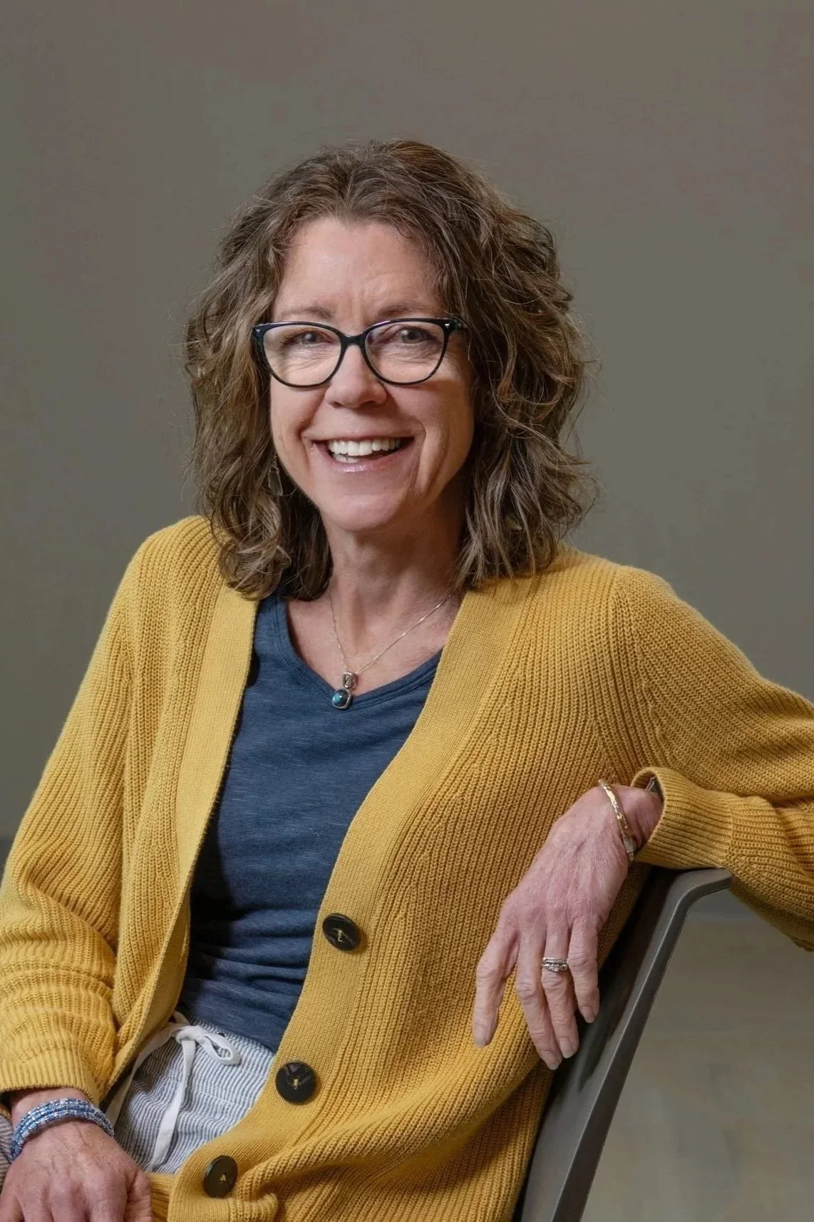 A woman with curly hair, glasses, and a yellow cardigan smiling while sitting on a chair.