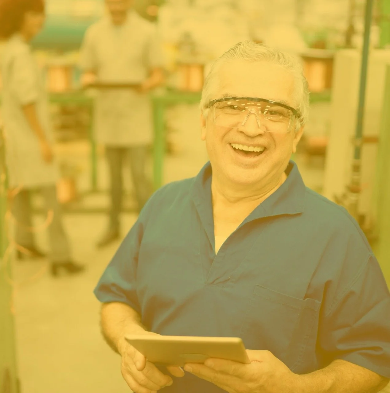 An older man wearing safety glasses and a blue shirt, smiling and holding a tablet, in a busy warehouse or manufacturing environment with workers in the background.