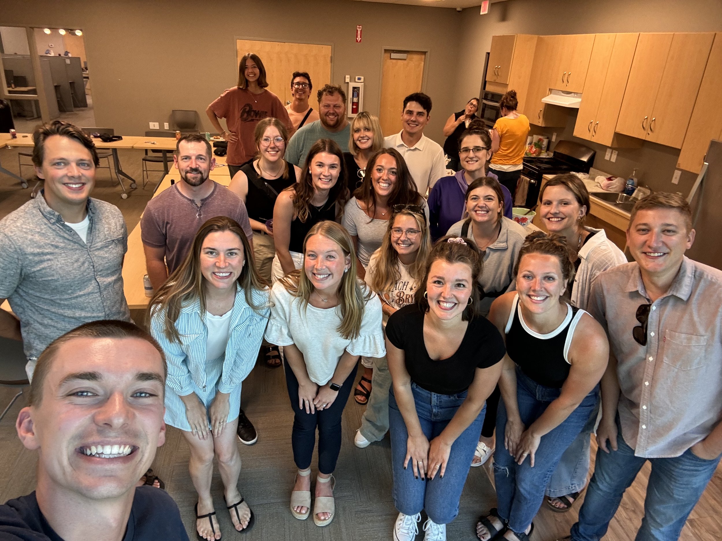 Group of people smiling and posing for a photo in a room with wooden cabinets and a kitchen area in the background.