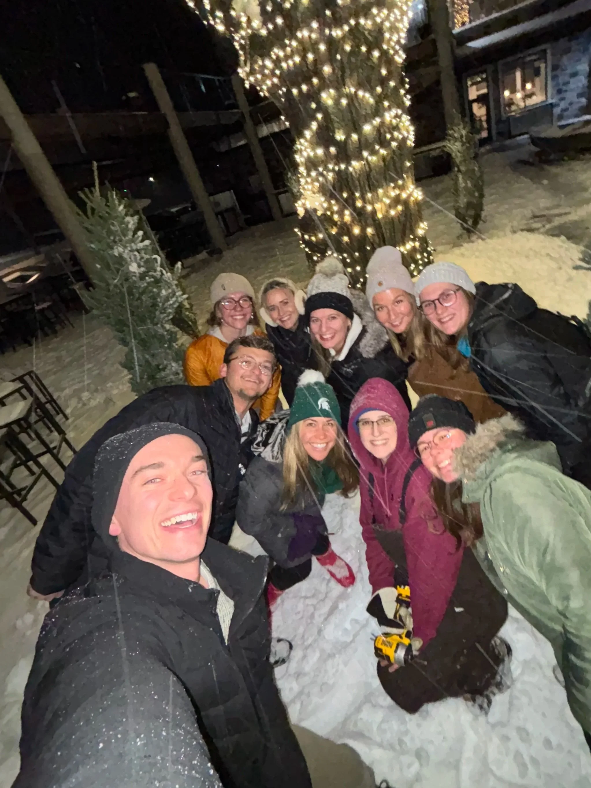 Group of people dressed warmly in winter clothing, smiling and taking a selfie outdoors at night during a snowy evening. Behind them, a large tree decorated with string lights and snow-covered evergreen trees are visible.