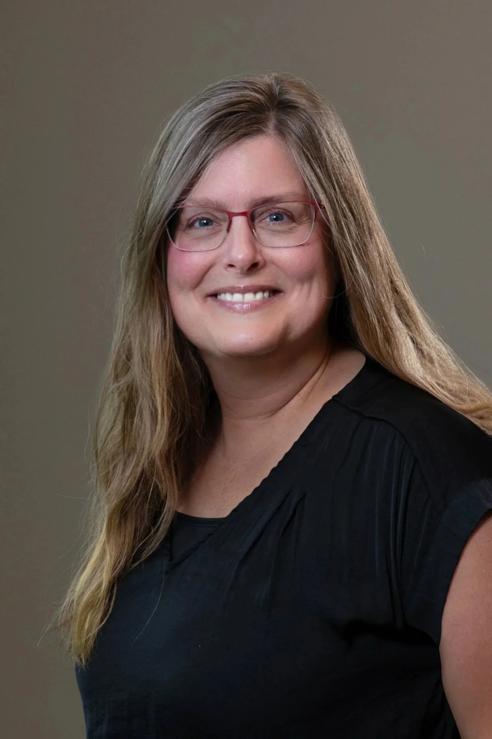 A woman with long, wavy brown hair and red glasses smiling at the camera, wearing a black top, in front of a plain gray background.