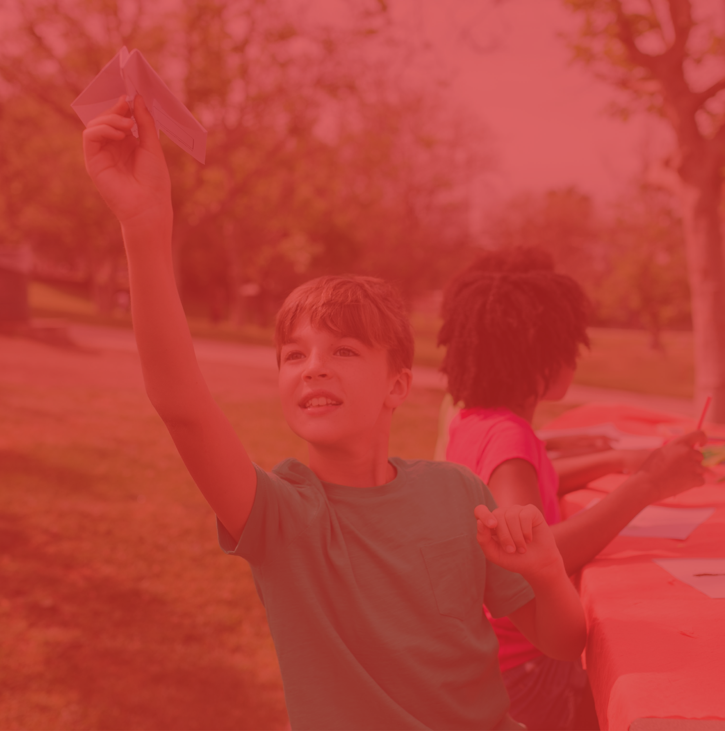 A young boy with brown hair smiling and raising his hand, holding a piece of paper, outdoors with trees and a table in the background.