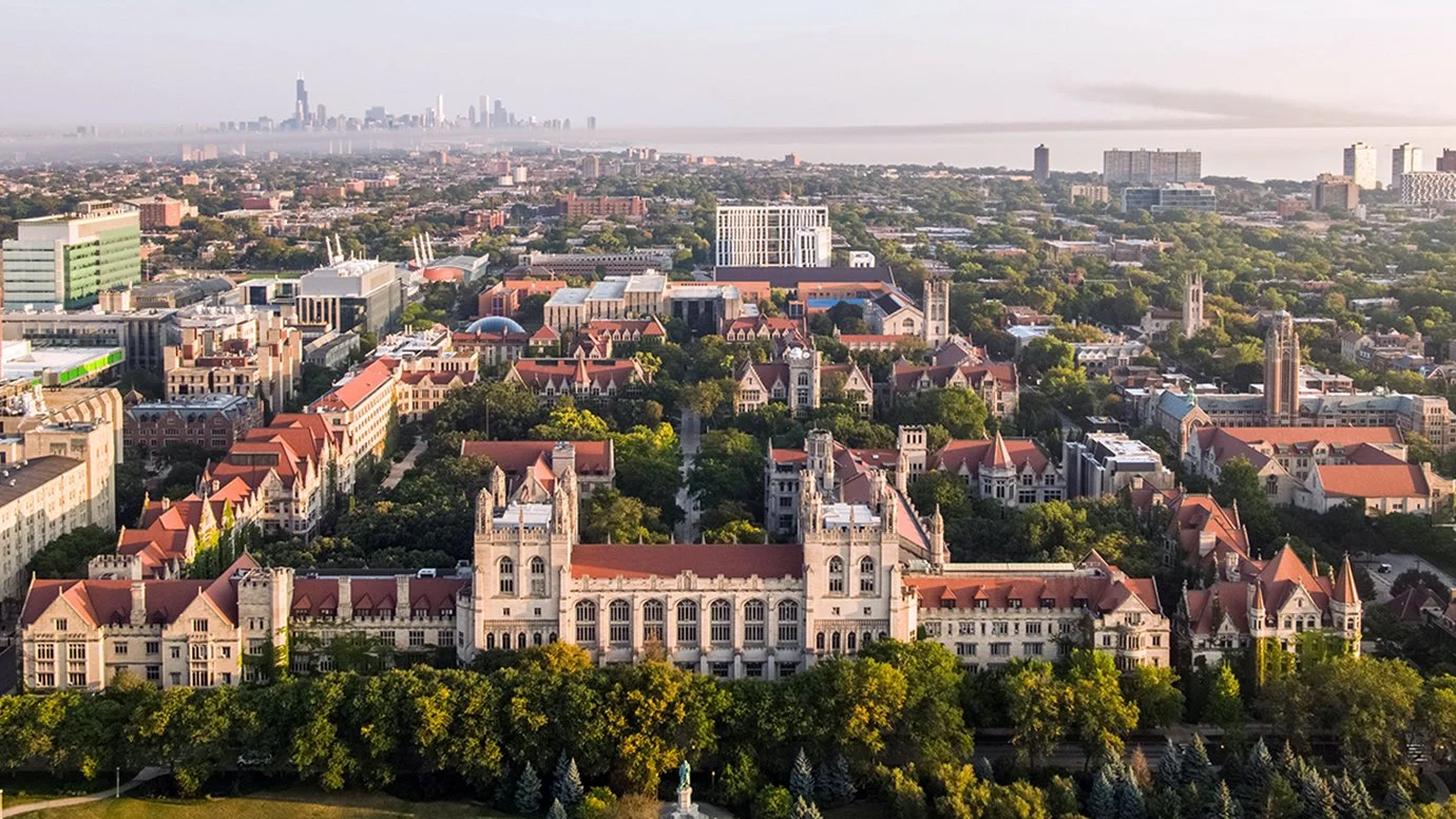 Aerial view of a historic university campus with Gothic architecture, red roofs, and lush green trees, with a modern city skyline and water in the background.