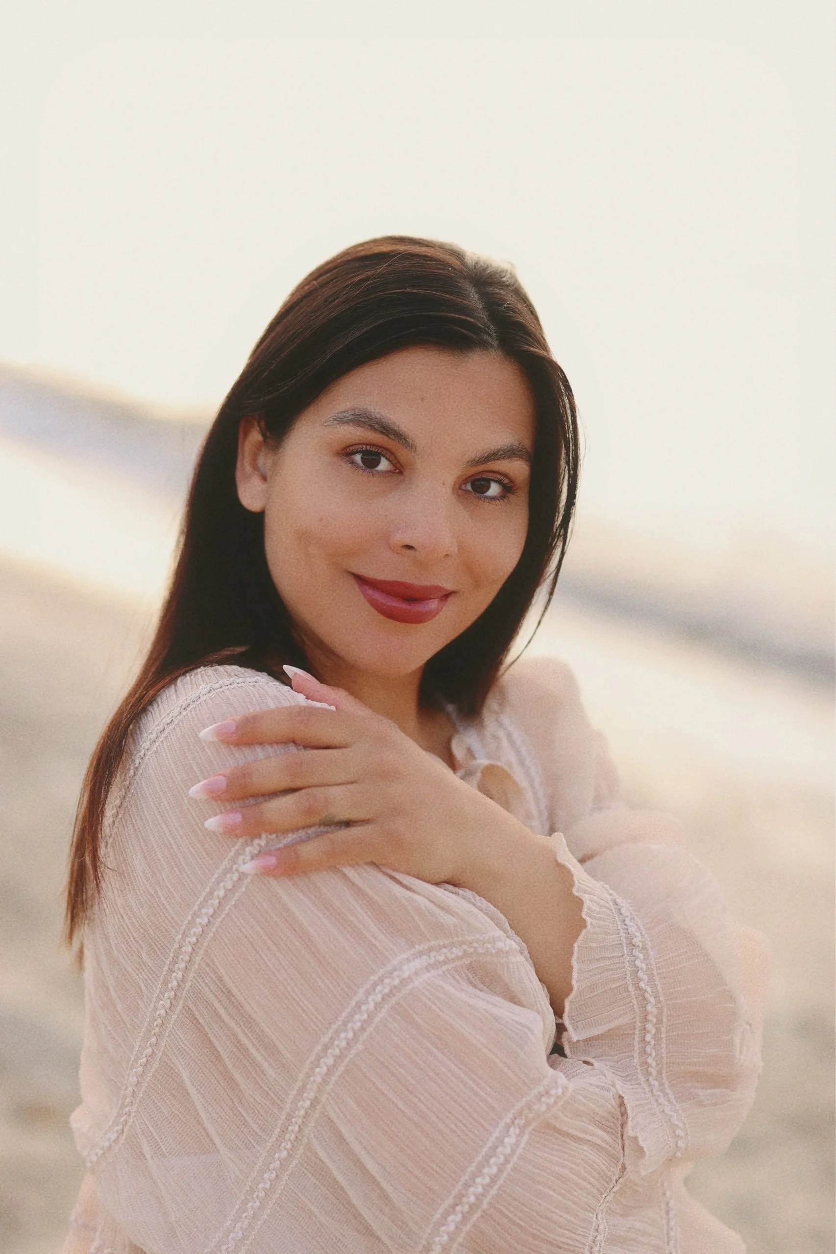 Junge Frau mit langen braunen Haaren, was für einen lichten Look sorgt, posiert am Strand bei Sonnenuntergang, trägt eine helleBluse