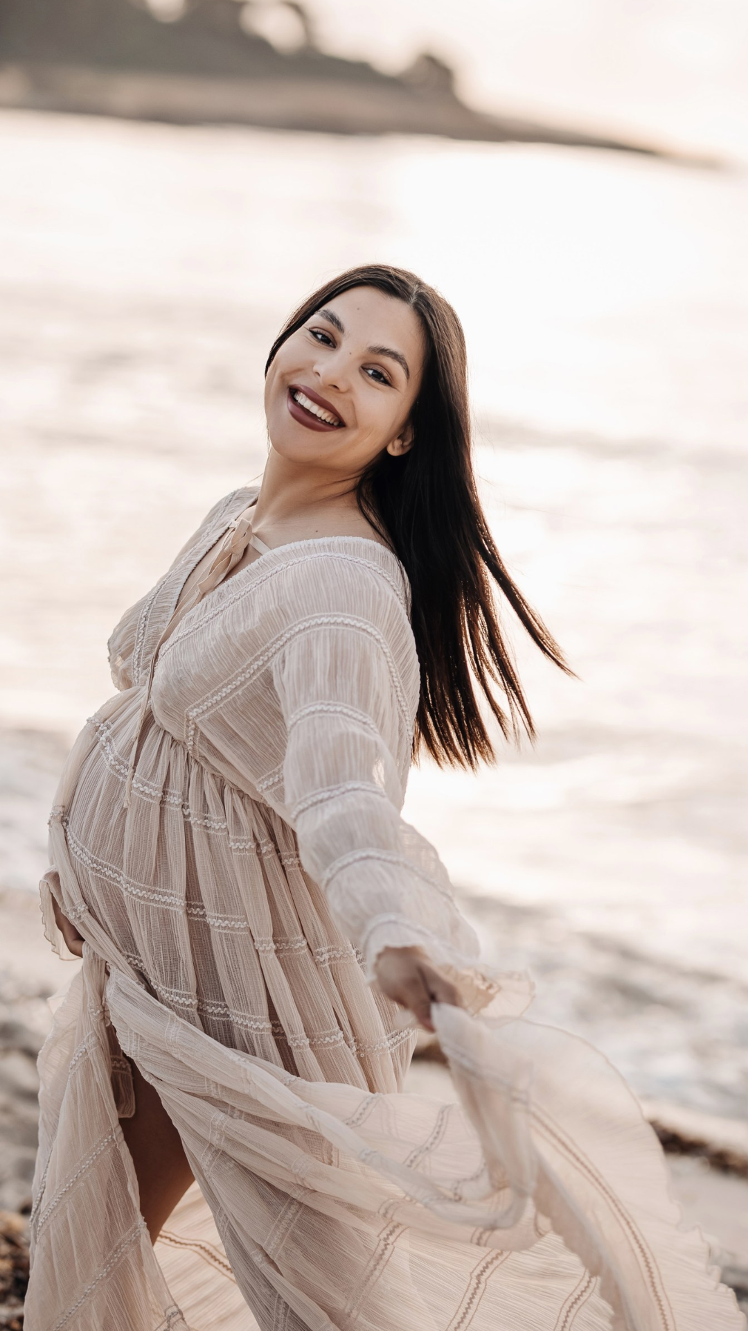 Lächelnde schwangere Frau bei Sonnenuntergang am Meer, trägt ein leichtes, langes Kleid.