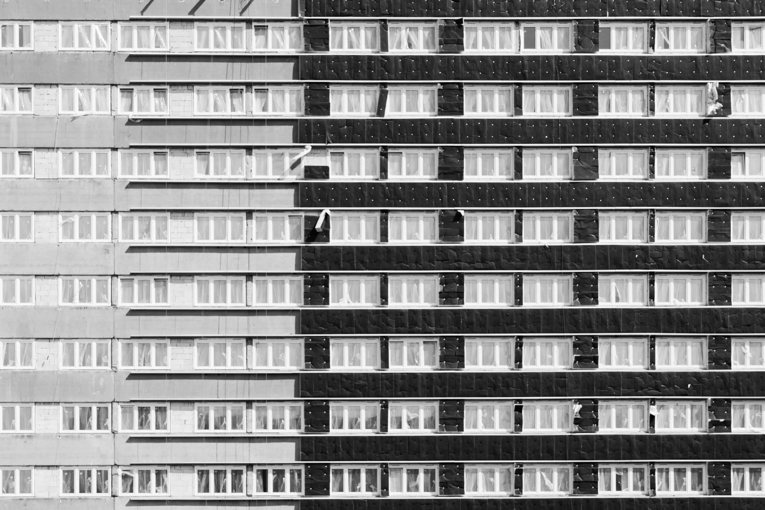 Black and white photo of the facade of a multi-story apartment building with rows of windows and balconies, some with open windows or curtains, and window cleaning equipment hanging from some of the windows.