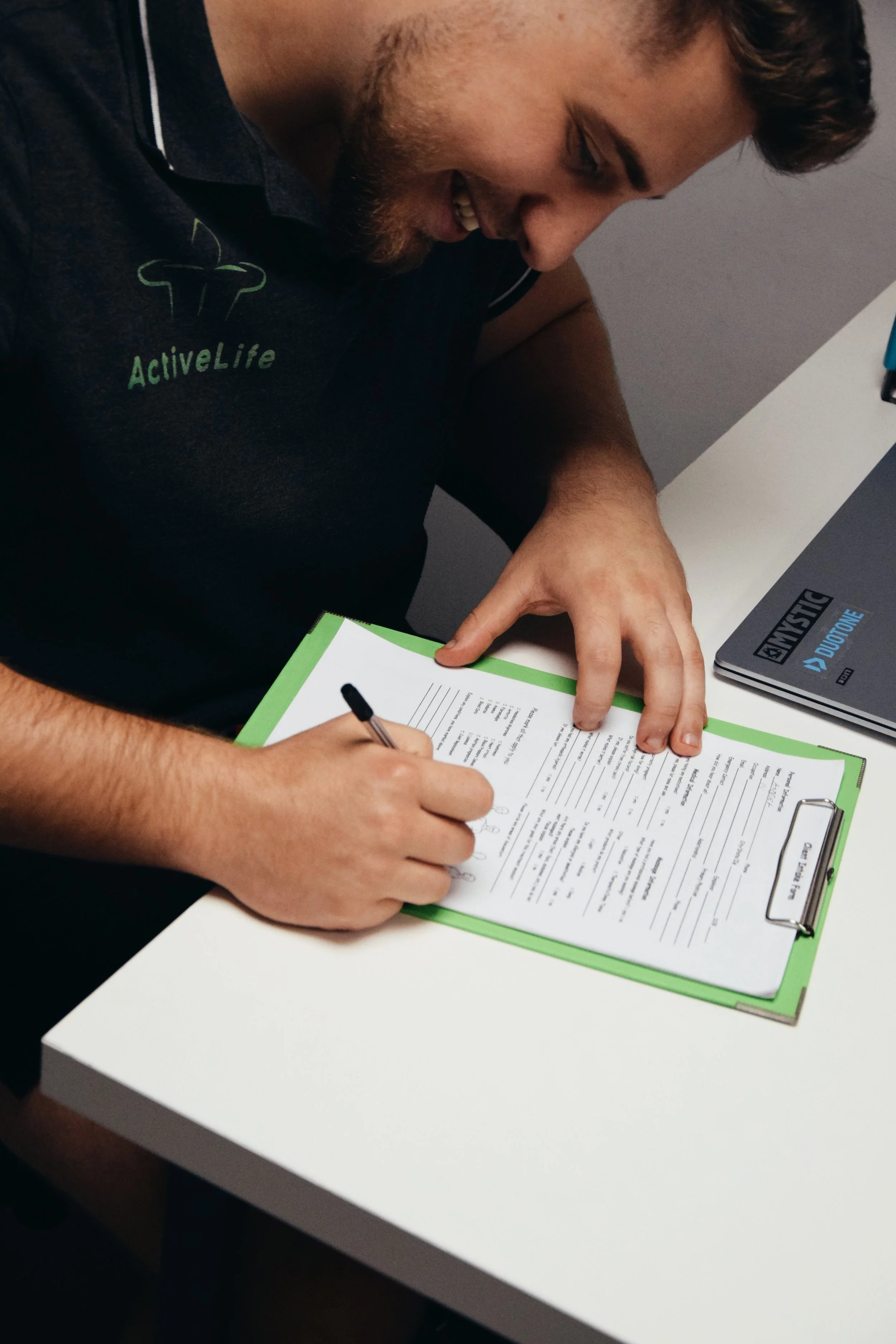 A man in a black shirt sitting at a white desk, filling out a form on a green clipboard with a black pen, smiling as he writes.