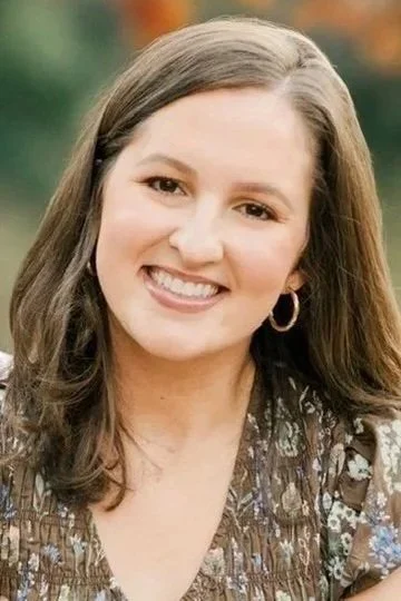 A young woman with brown hair, smiling, wearing hoop earrings and a patterned top, outdoors.