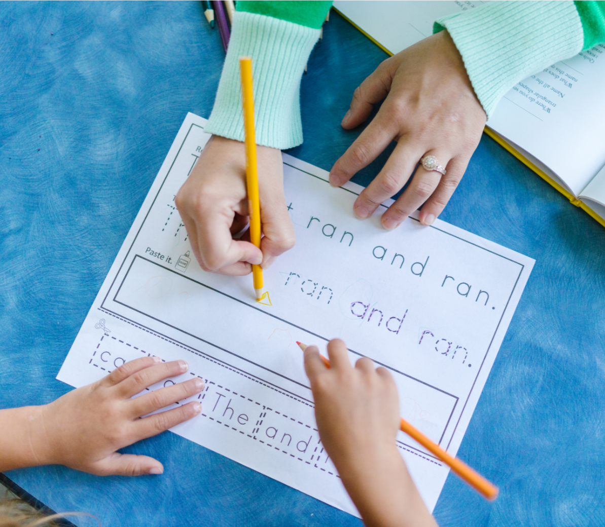 A child's and an adult's hands working on a handwriting worksheet about the words 'ran' and 'and' on a blue table, with colored pencils and a book nearby.