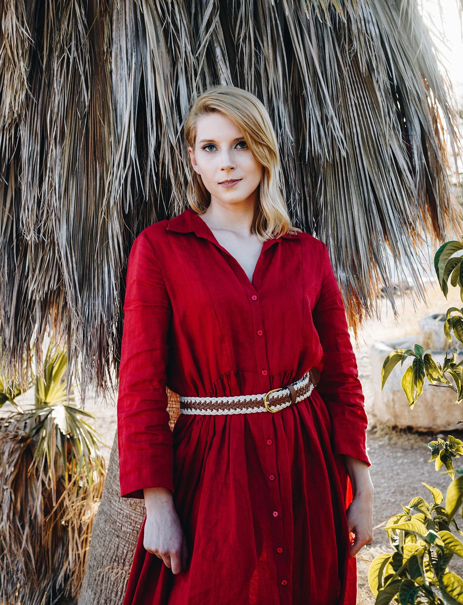 A woman with blonde hair wearing a red dress and a woven belt, standing outdoors in front of a thatched tiki hut with tropical plants around her.