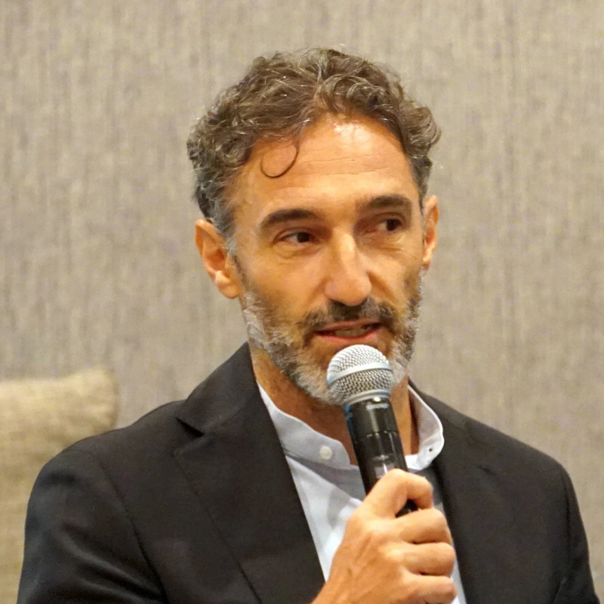 A middle-aged man with curly gray hair and a beard, dressed in a black suit and white shirt, speaking into a microphone. He is indoors against a neutral beige background.