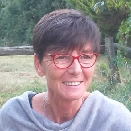 A smiling woman with short brown hair and red glasses outdoors in a natural setting.