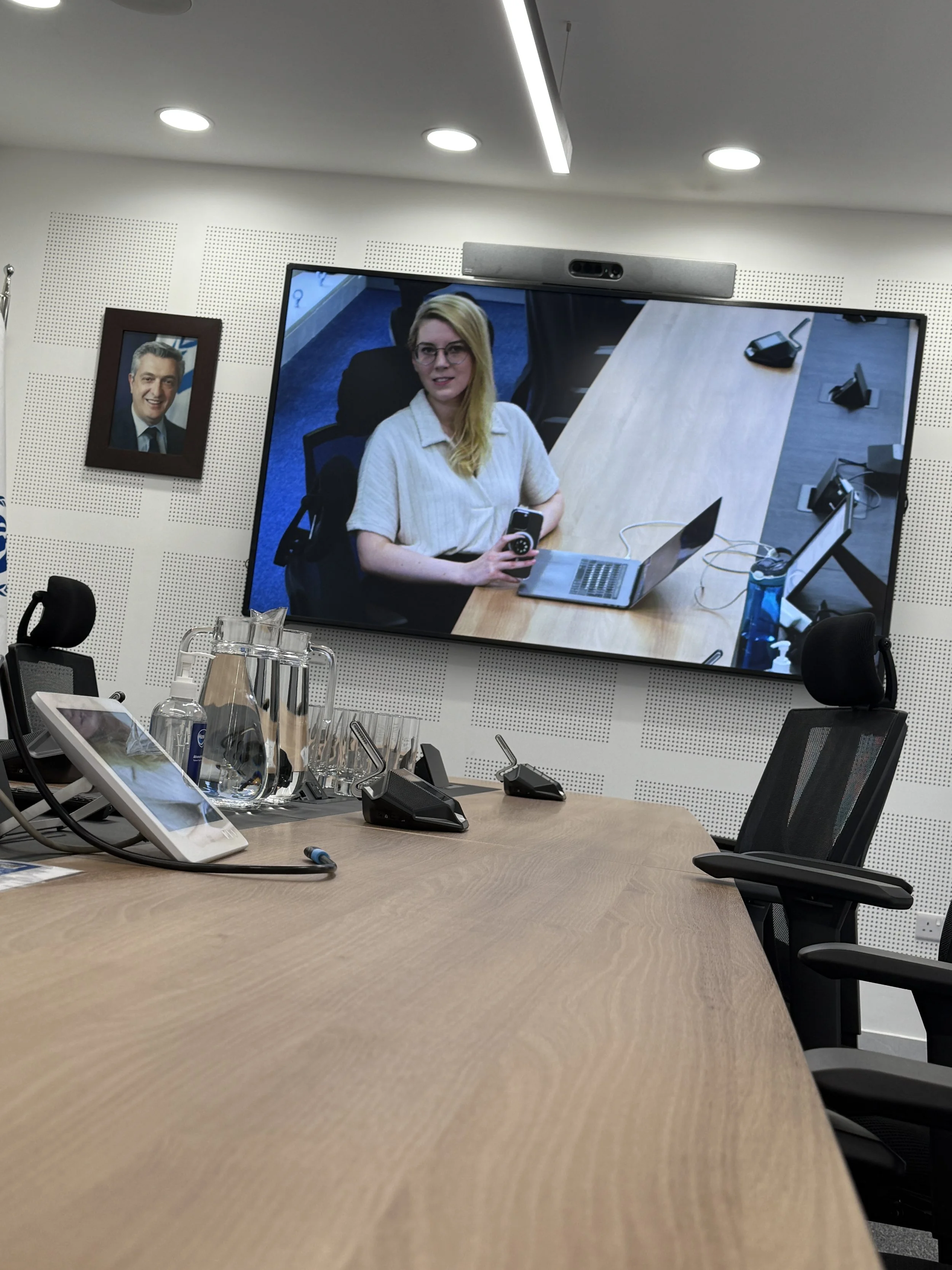 Video conference room with a large screen displaying a woman in a white shirt holding a phone, a portrait of a man on the wall, and conference tables with phones and water bottles.