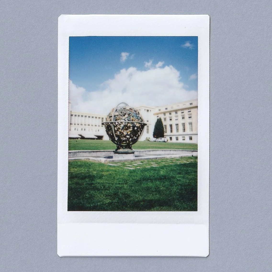 A sculpture of a globe with world map design in front of a large white building with multiple windows, on a grassy area under a blue sky with clouds.
