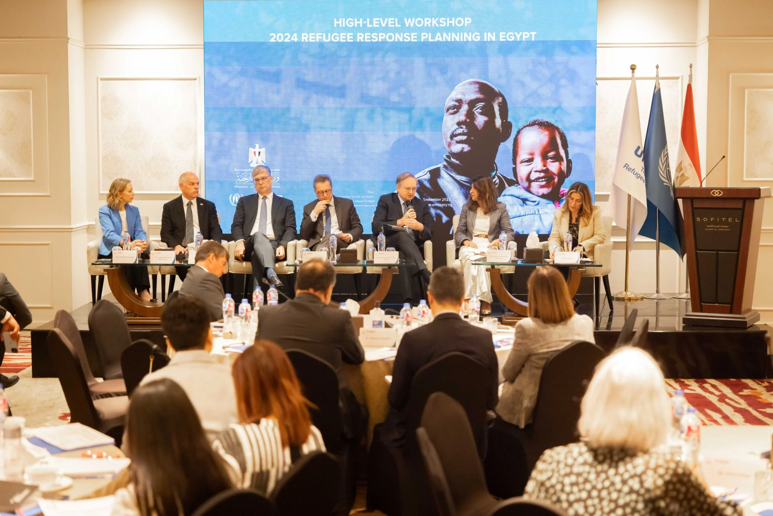 A group of six people seated on stage at a panel discussion during a workshop on refugee response planning in Egypt, with a large screen behind them displaying the workshop topic and images of two children. The setting is a conference room with flags to the side and an audience in front.