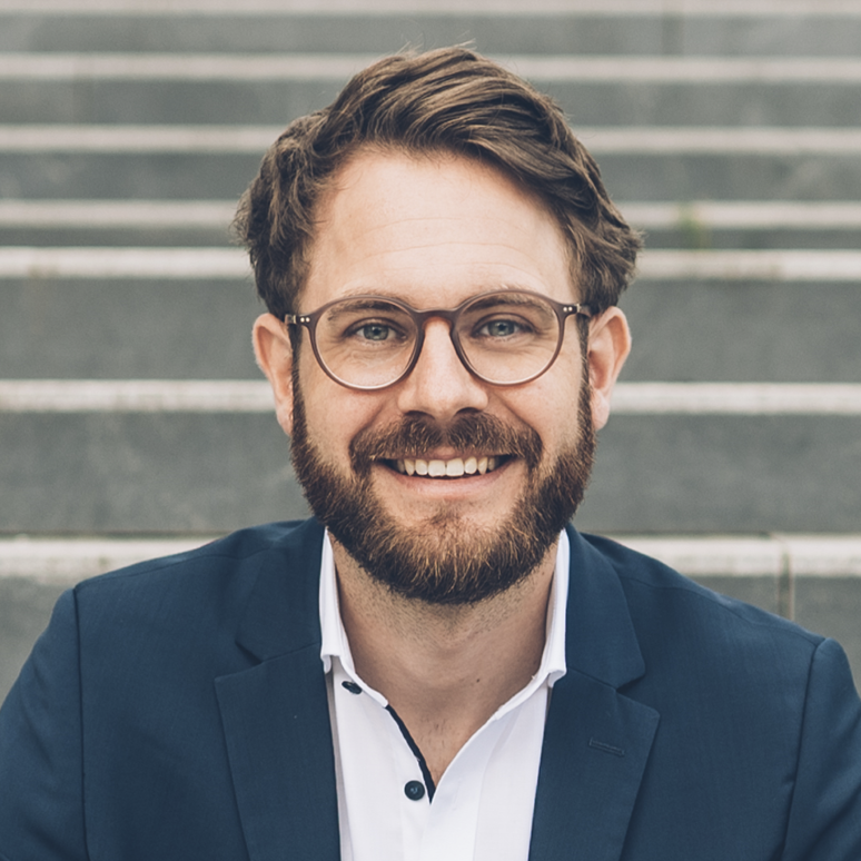 Smiling man with glasses and beard in a business suit, sitting on outdoor stairs.