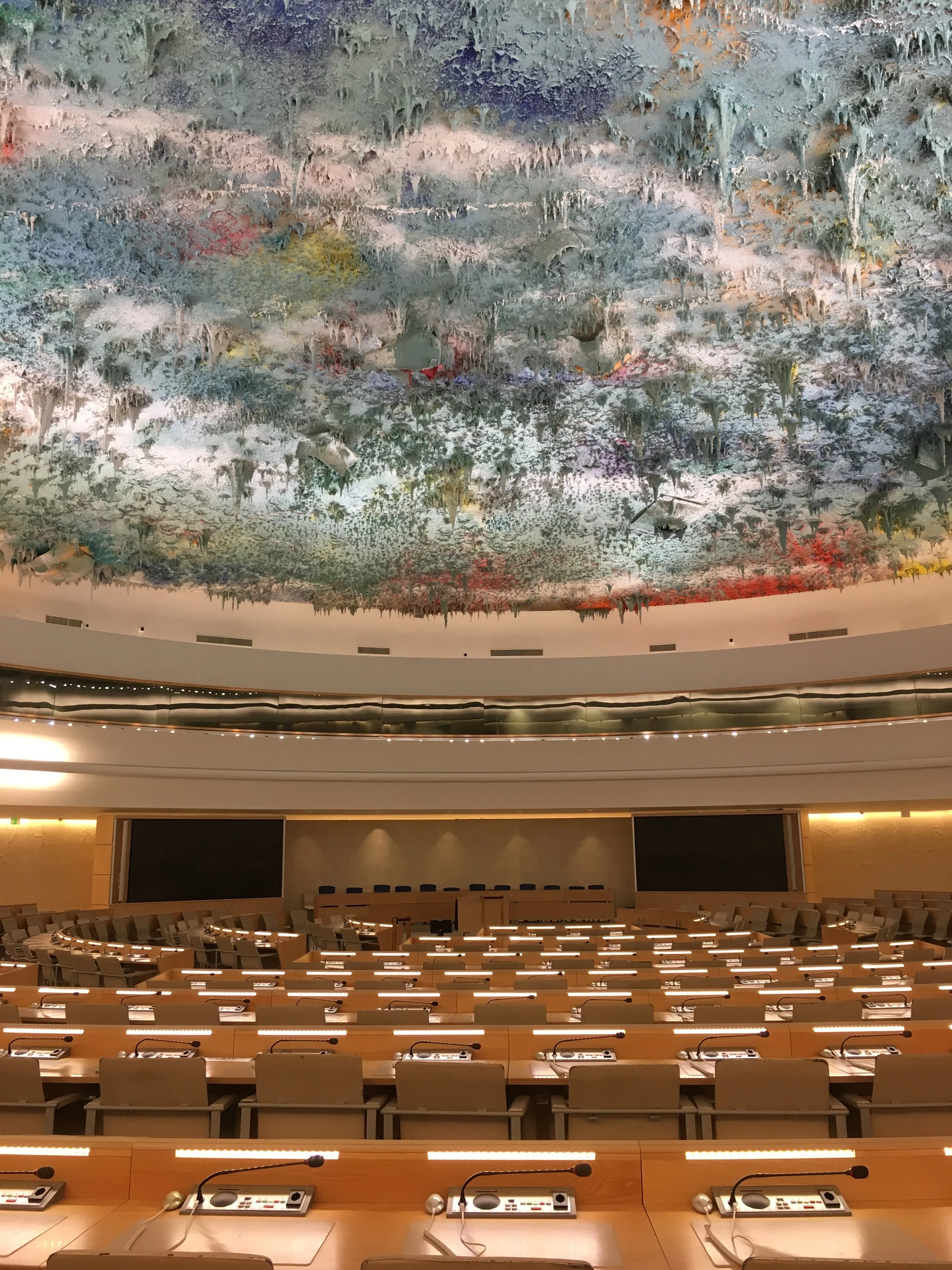 The image shows a large, empty conference hall with rows of wooden desks and chairs, each equipped with a microphone and electronic voting devices. There is a balcony area with a curved design and a ceiling mural of an atmospheric, abstract sky with clouds, trees, and color blending.