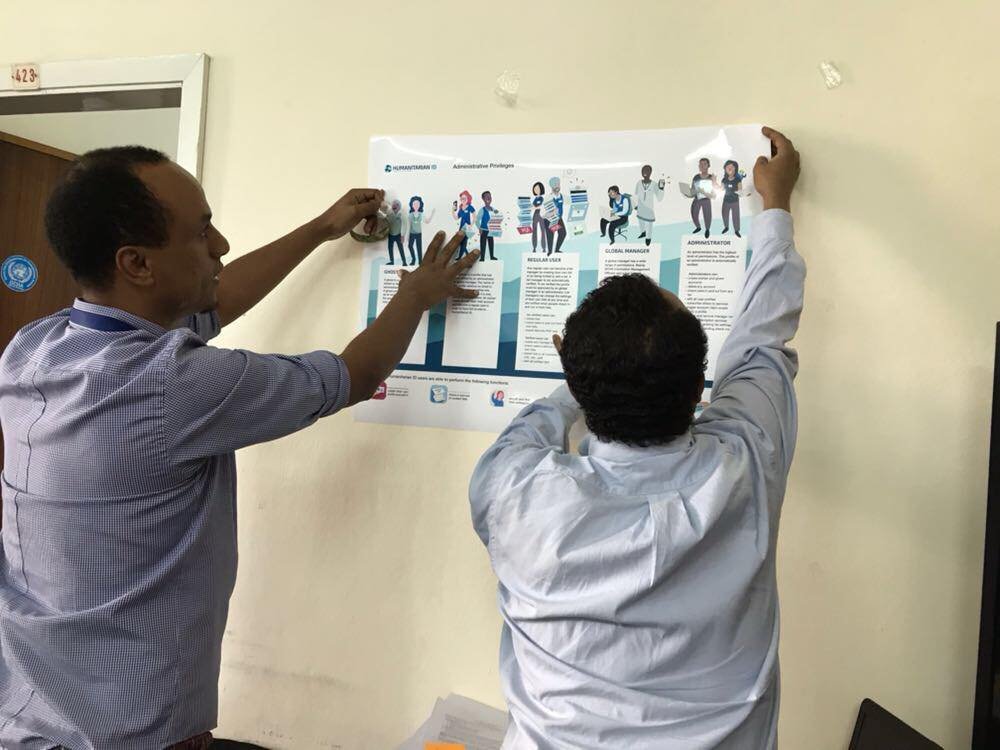 Two men hang a large informational poster featuring illustrated characters and text on a beige wall.