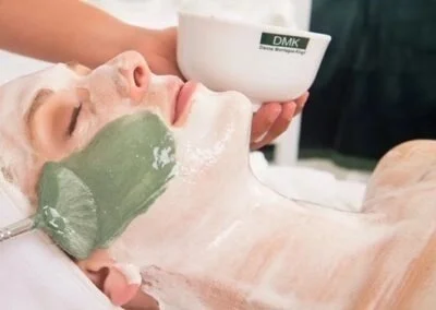 A woman receiving a facial treatment with a green mask, lying down with closed eyes, while a person applies a white substance to her face and holds a bowl labeled 'DMK'.