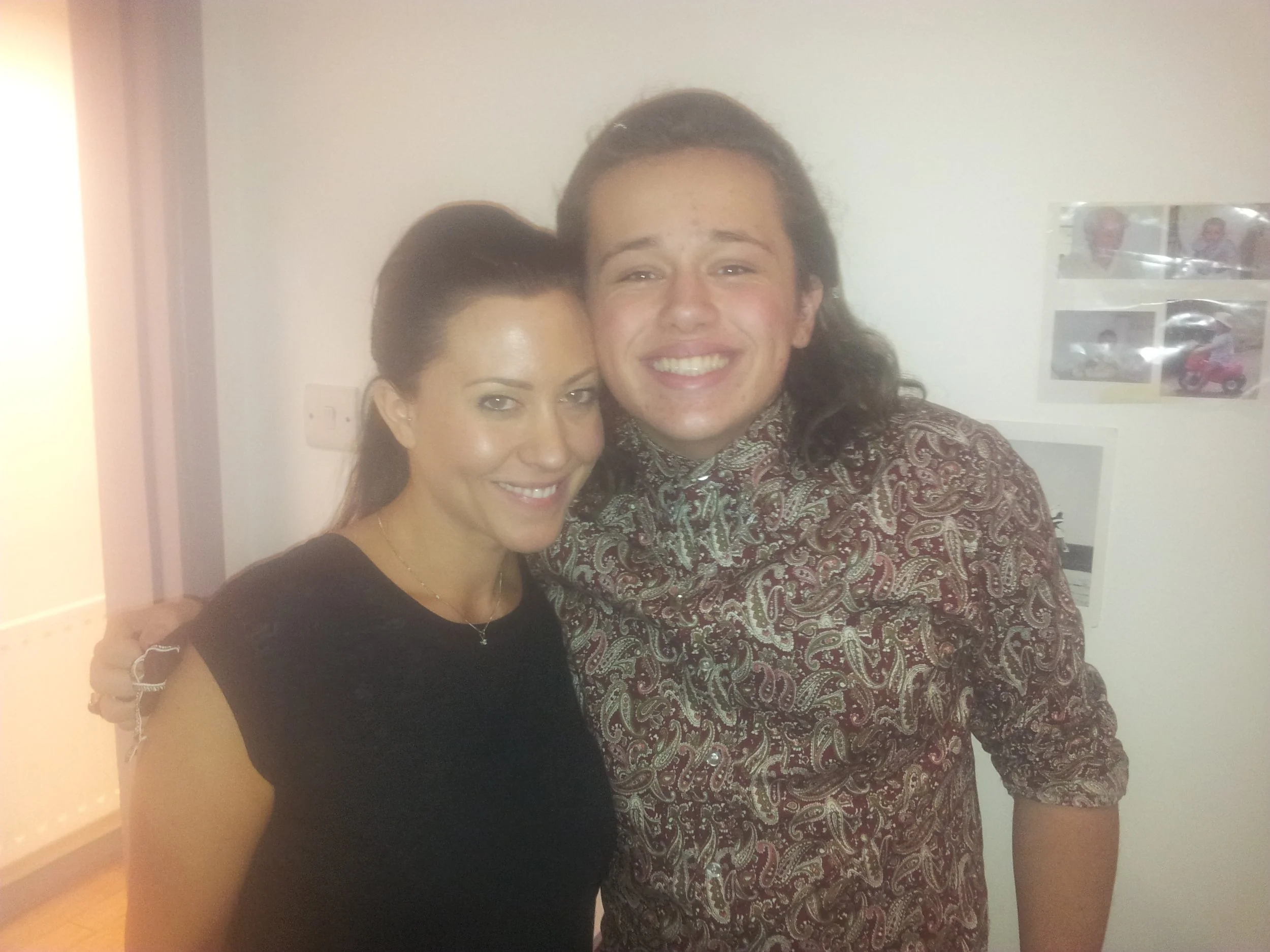 A young man and woman smiling and posing together indoors, with a white wall and some photographs in the background.