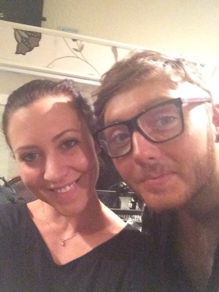 A woman and a man taking a close-up selfie together indoors. The woman has dark hair pulled back, fair skin, and is smiling. The man is wearing glasses, has light skin, and a slight smile.