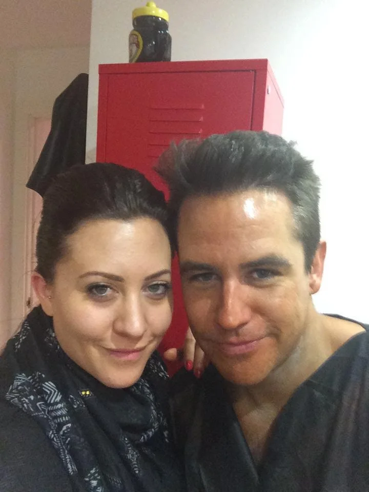 Two people smiling and taking a selfie indoors, with a red cabinet and a black and yellow water bottle on top in the background.