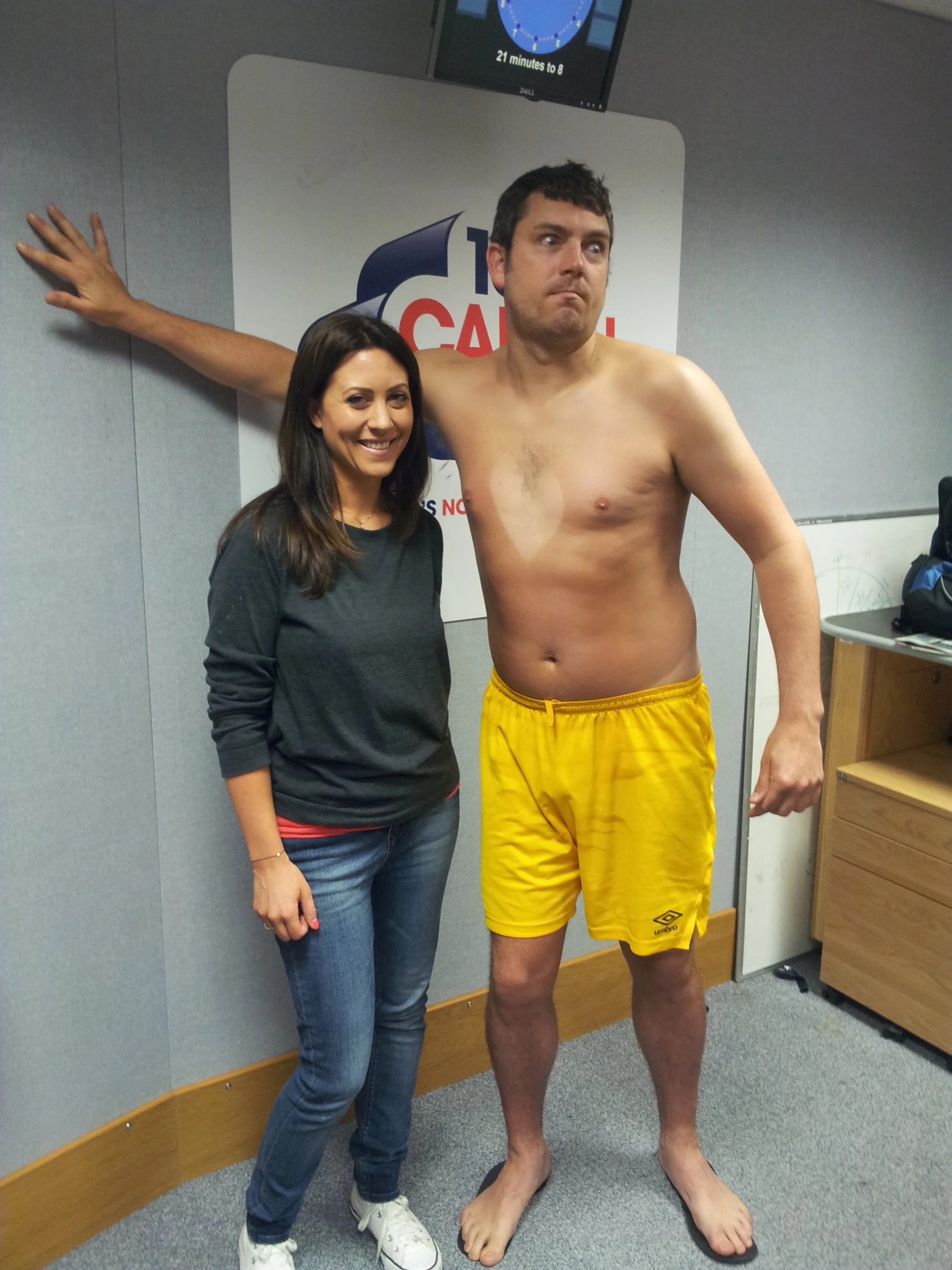A woman and a shirtless man standing together in a room with gray walls, a clock, and a sign, smiling and posing for a photo.