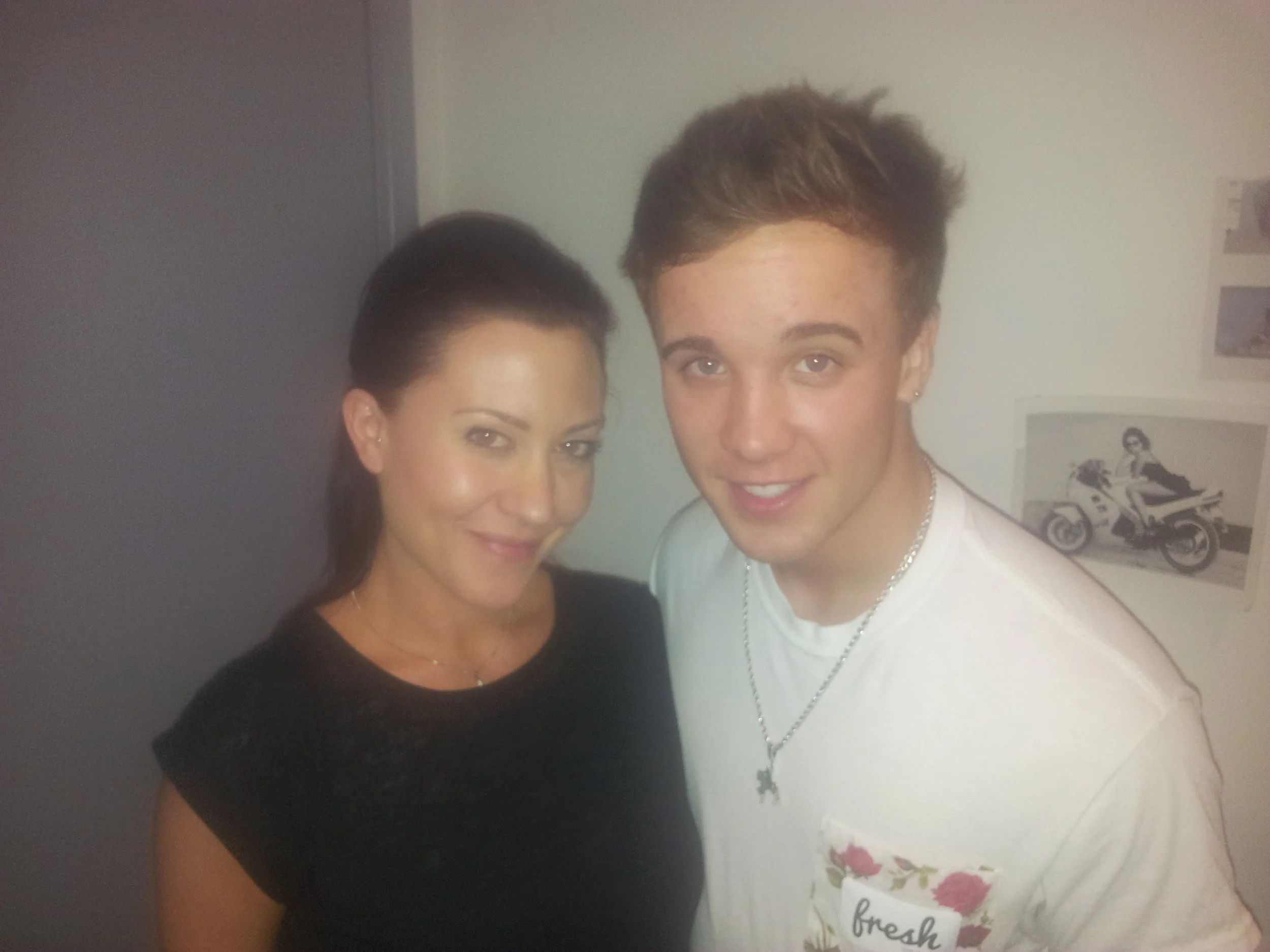 A woman with dark hair and a man with light brown hair smiling, standing close together indoors, with framed pictures on the wall behind them.
