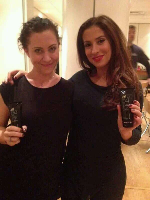 Two women standing close together in black outfits, each holding a small black tube of skincare or cosmetic product, in an indoor setting.